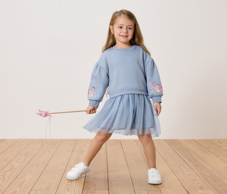 Virágmintás hímzéssel díszített kék pulóvert és tüllszoknyát viselő kislány, kezében varázspálcát tart.
