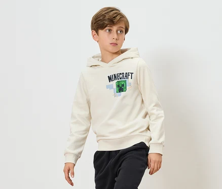 Egy fiú krémszínű Minecraft kapucnis pulóvert és fekete melegítőalsót visel fehér háttér előtt.
