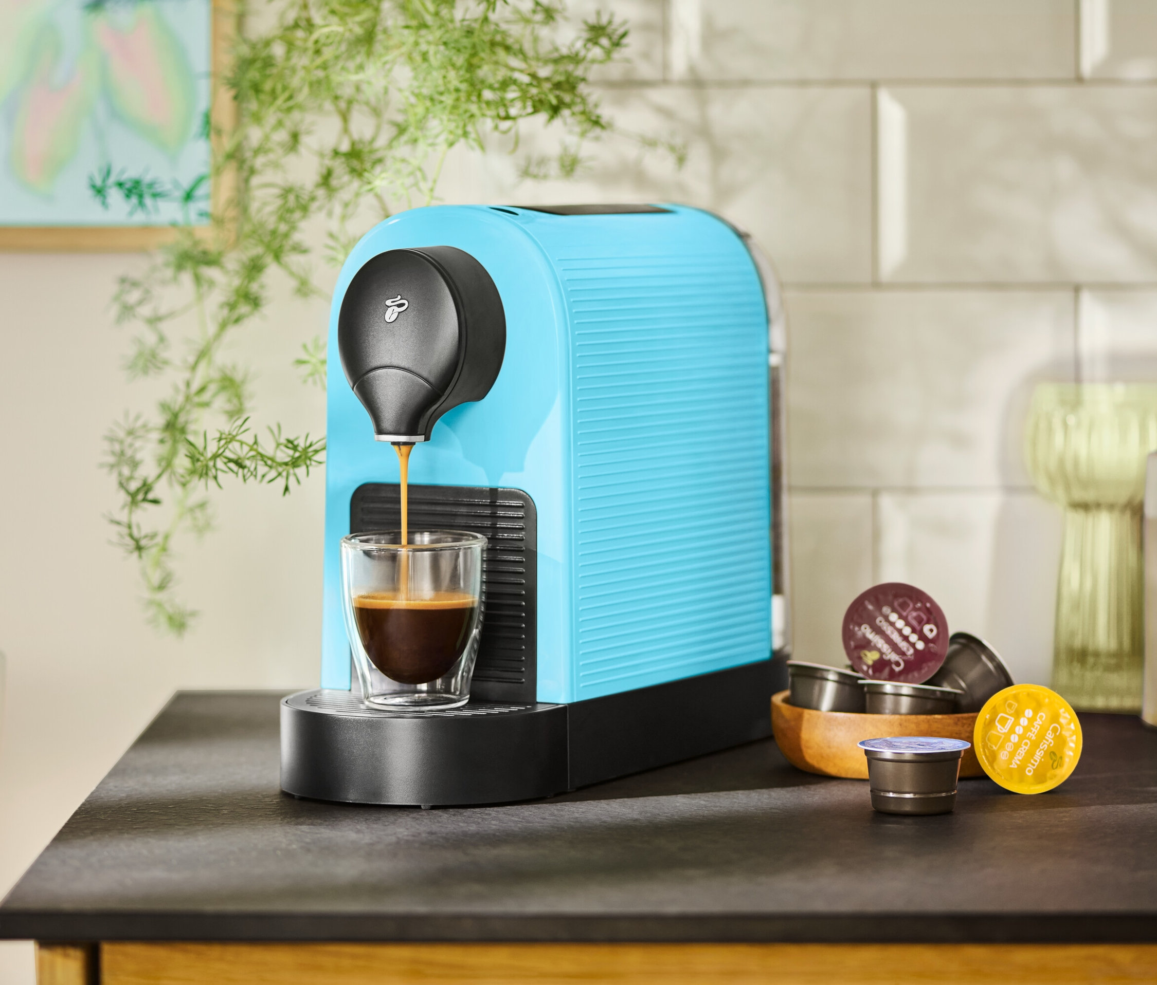 Egy türkizkék kávéfőző kávéval tölt meg egy csészét. Mellette egy fából készült tálban kávékapszulák találhatók.