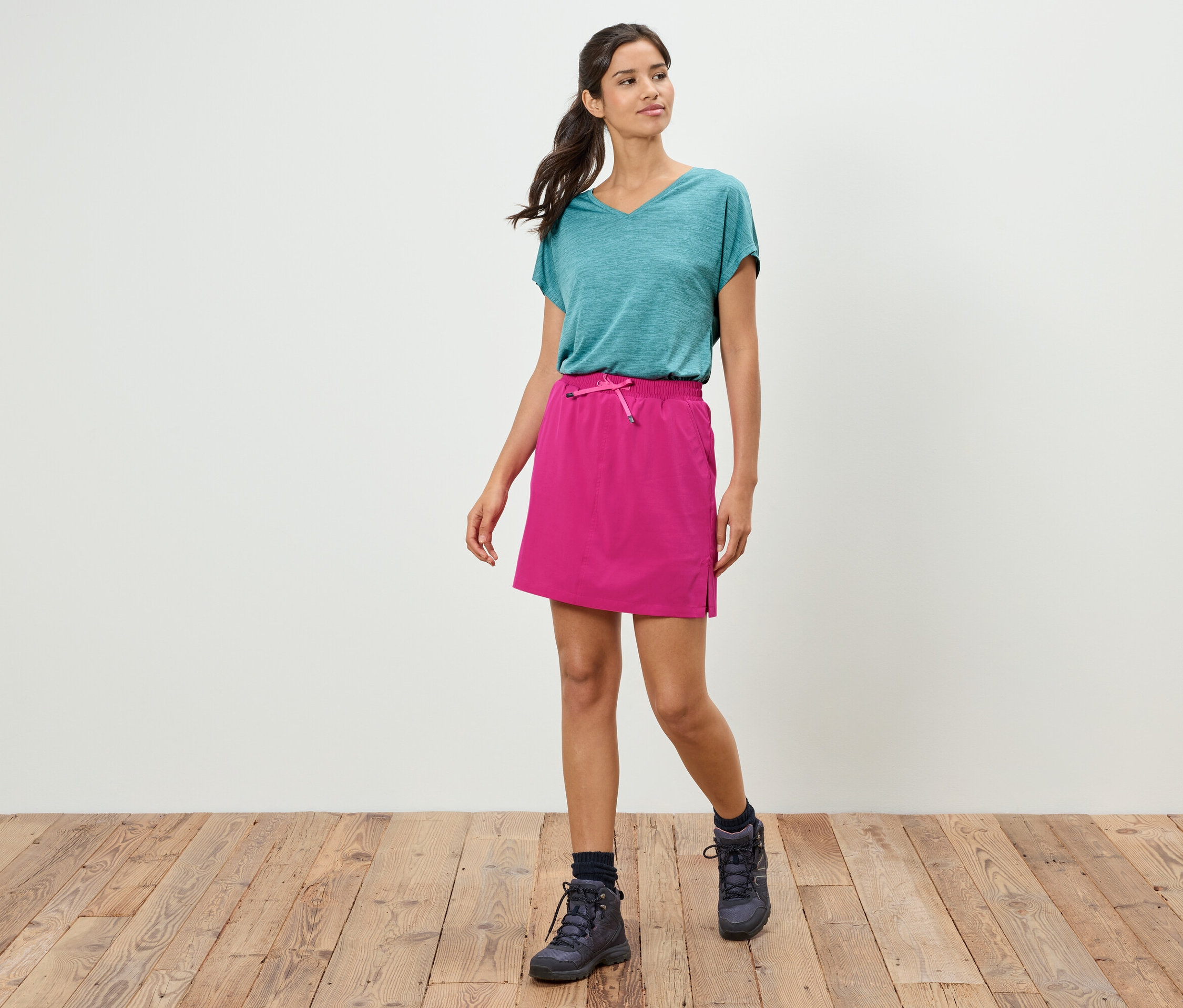 Nő sétál türkizkék pólóban, rózsaszín szoknyában és túrabakancsban a fapadlón egy fehér fal előtt.