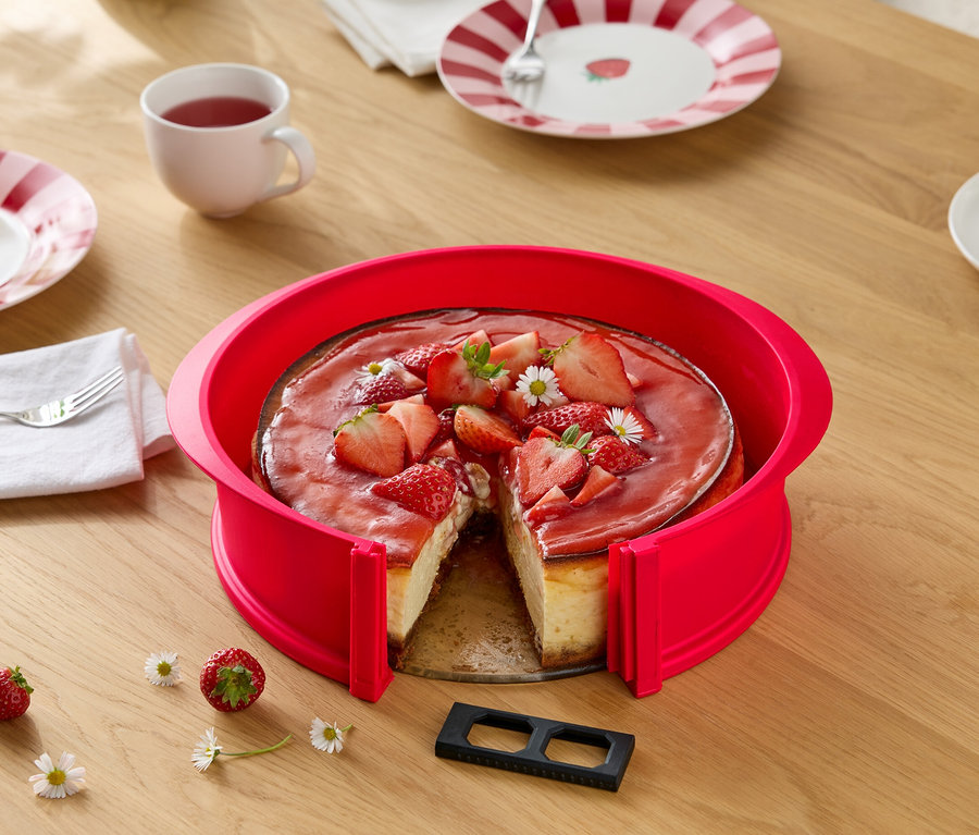 Epres sajttorta piros tortakarikával, szeletelve, fa asztalon százszorszépekkel, terítékkel és italokkal.