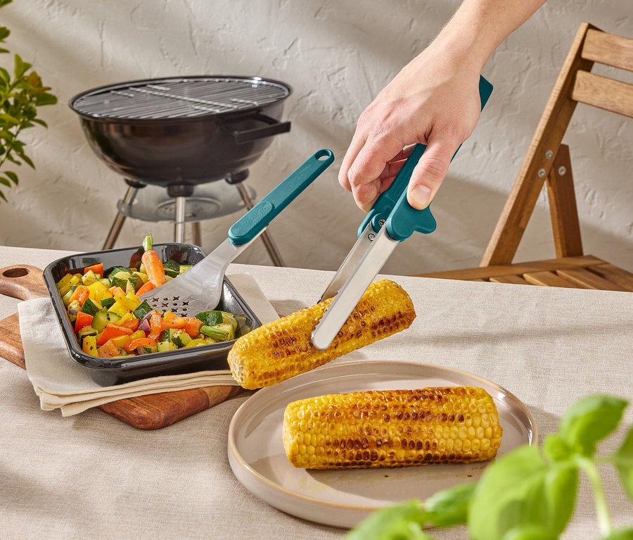 Egy személy grillezőcsipesszel egy kukoricacsövet tart egy tányér fölött. Az asztalon zöldségek és egy grill is található.