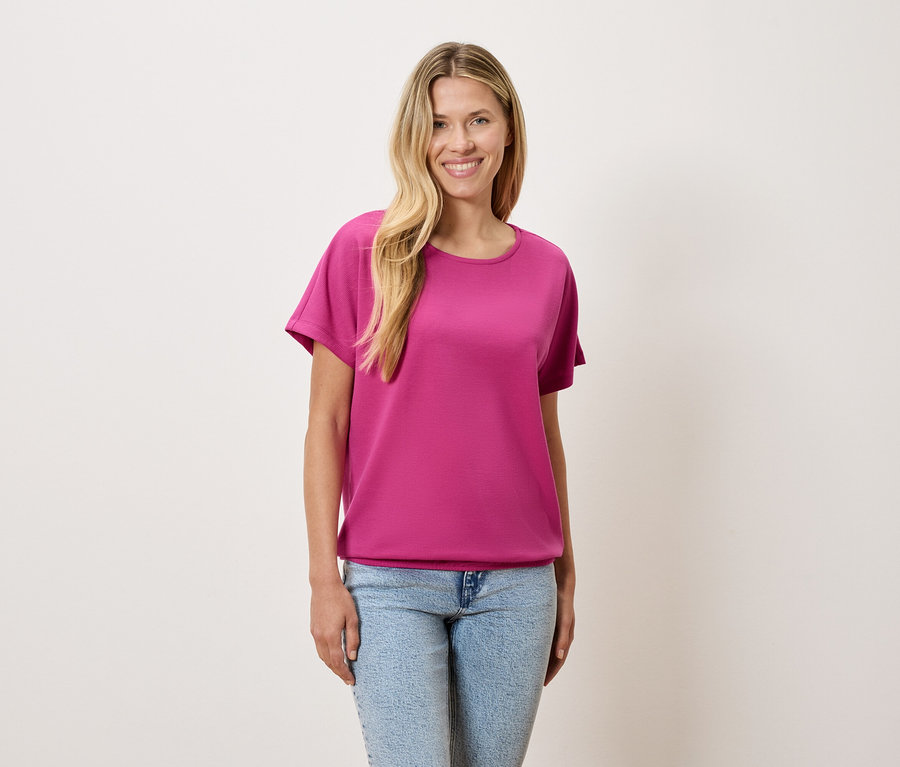 Egy szőke hajú nő rózsaszín pólót és kék farmert visel.