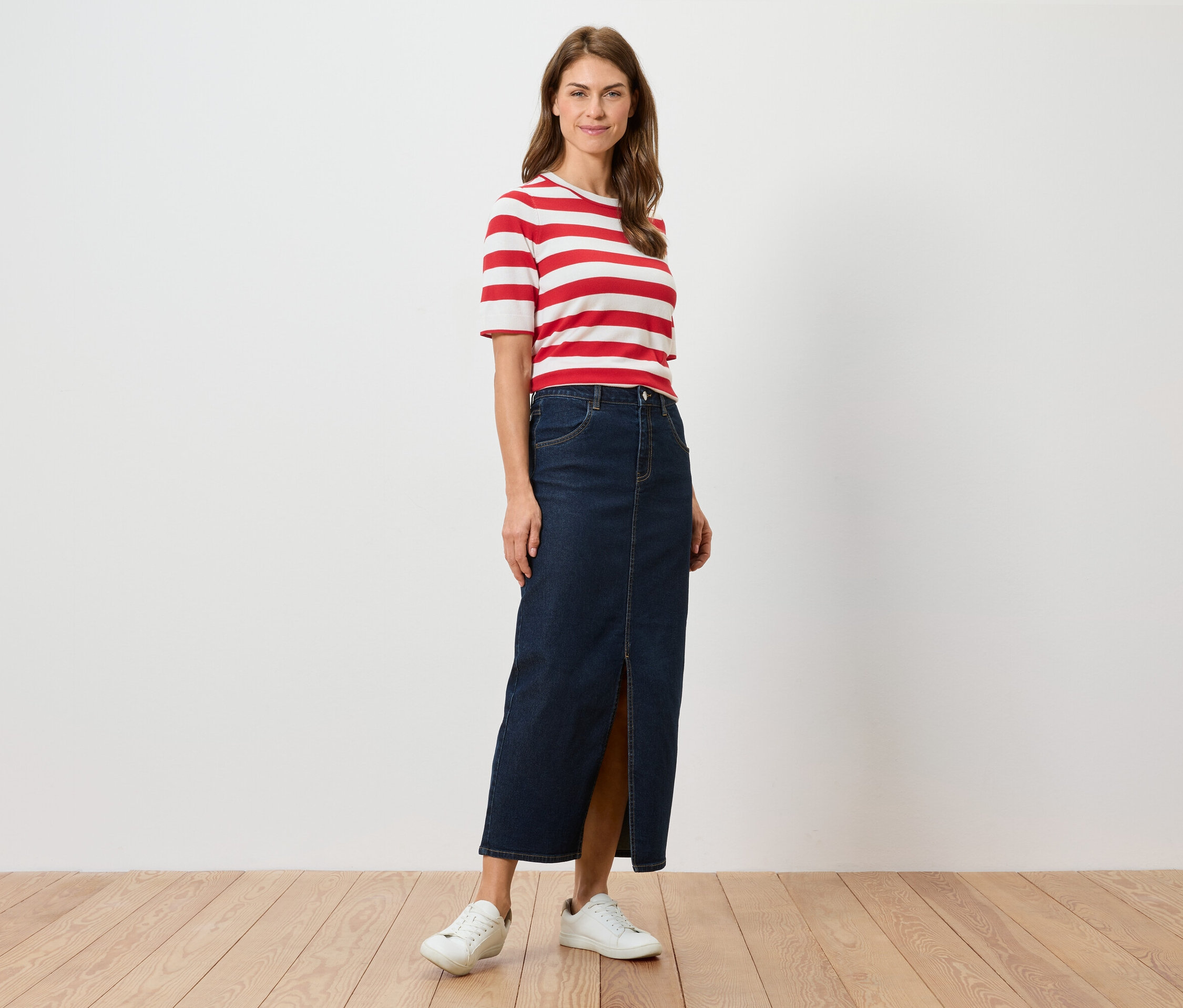 Egy nő áll, piros-fehér csíkos pólót, hosszú, sliccelt farmer szoknyát és fehér tornacipőt visel.