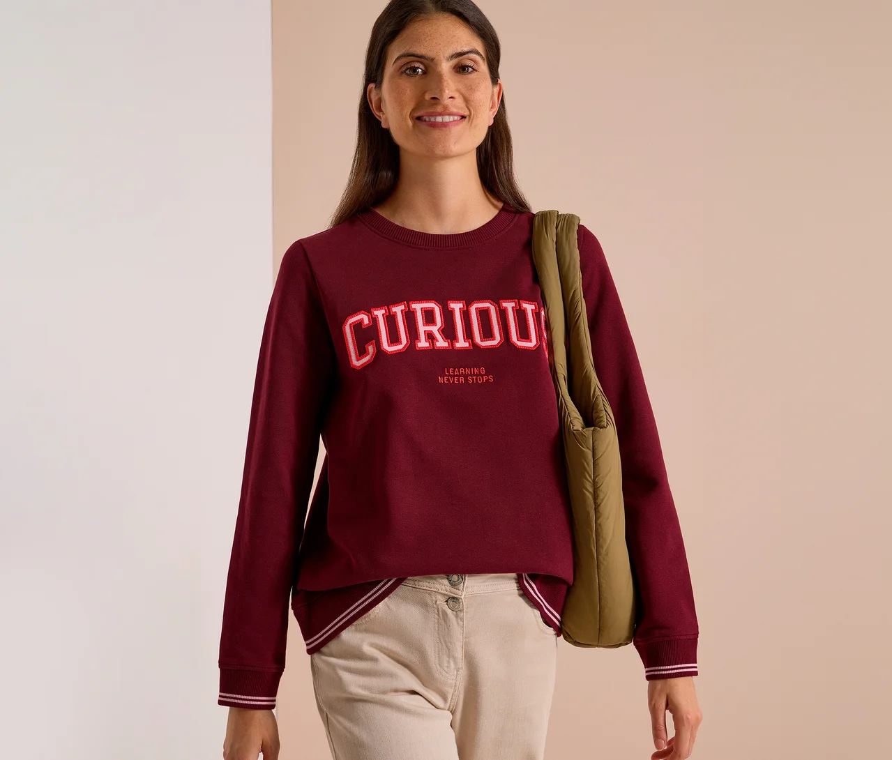 Mosolygó nő bordó CECIL szabadidőfelsőben, "CURIOUS LEARNING NEVER STOPS" felirattal, bézs nadrágban, zöld táskával a vállán.