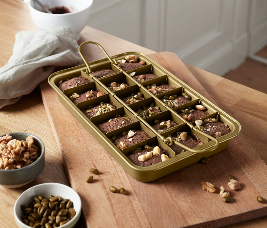 Egy arany színű, fogantyúval ellátott brownie sütőforma tele kis brownie-kkal egy vágódeszkán.