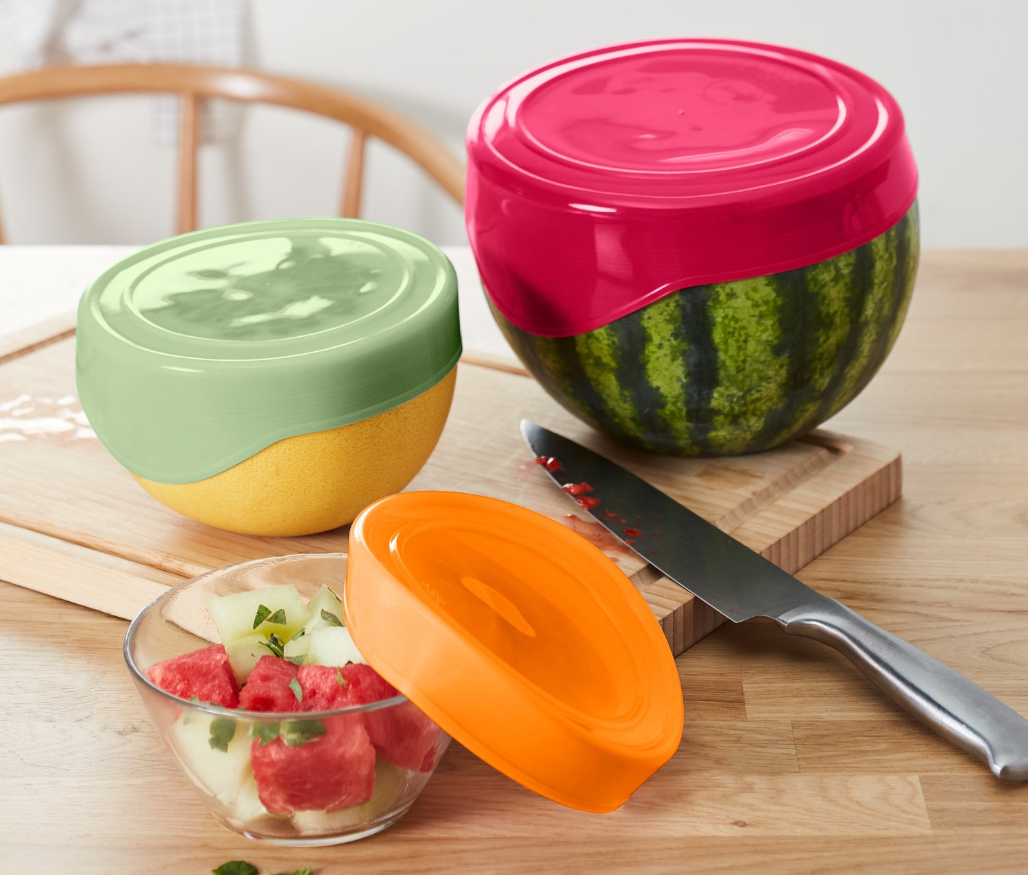 Egy fél görögdinnye és egy fél sárgadinnye 3 szilikon frissentartó fedéllel van letakarva. A görögdinnyesalátához tartozik egy narancssárga fedő és egy kés.