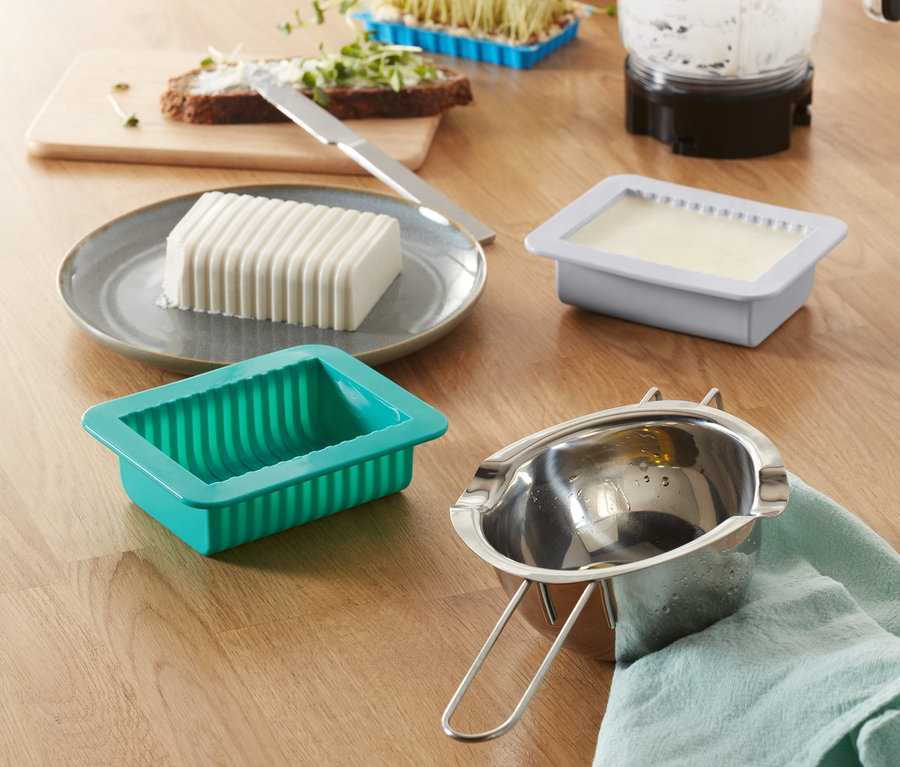 Egy asztalon egy növényi vaj készítő készlet, egy vajforma, egy turmixgép, hajtások és kenyér vajjal.