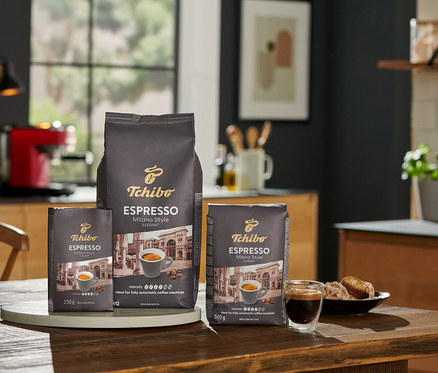 Három csomag Tchibo Espresso Milano Style kávébab 250 g, 500 g és 1 kg-os kiszerelésben egy fa asztalon a konyhában.
