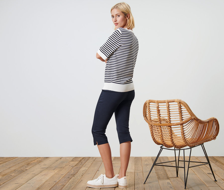 Nő pózol sötétkék 3/4-es sztreccsnadrágban és csíkos felsőben egy fonott szék mellett.