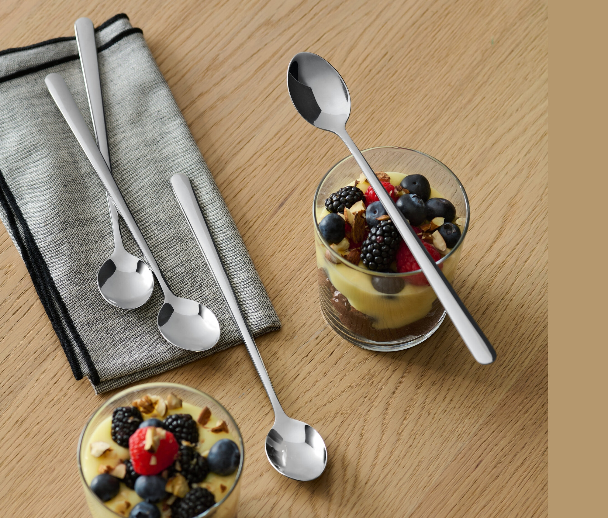 Egy fa asztalon egy szürke kendő és két pohár desszert és gyümölcs látható. Minden pohár mellett egy-egy 4 Latte Macchiato kanálból.