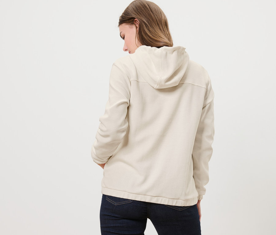 Hátulról látható egy nő, kezével a zsebében, krémszínű szabadidőfelsőt visel.