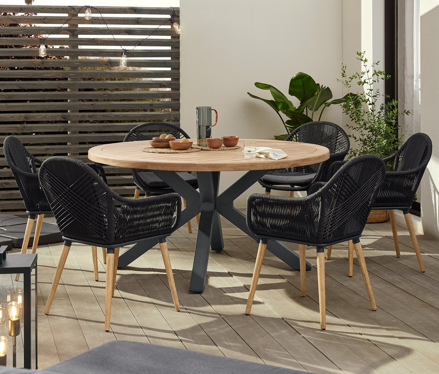 Terasz kerek teakfa asztallal, hat fonott székkel körülvéve, LED-es égősorral megvilágítva.