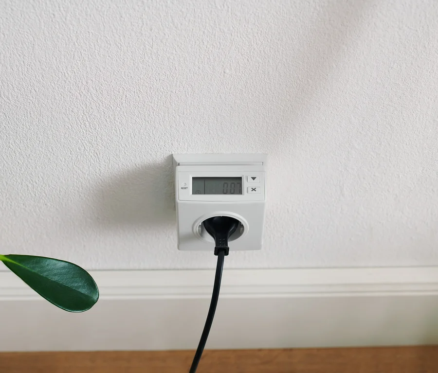 Fehér energiafogyasztás-mérő egy fehér fali aljzatba dugva, fehér falon, fekete kábellel.