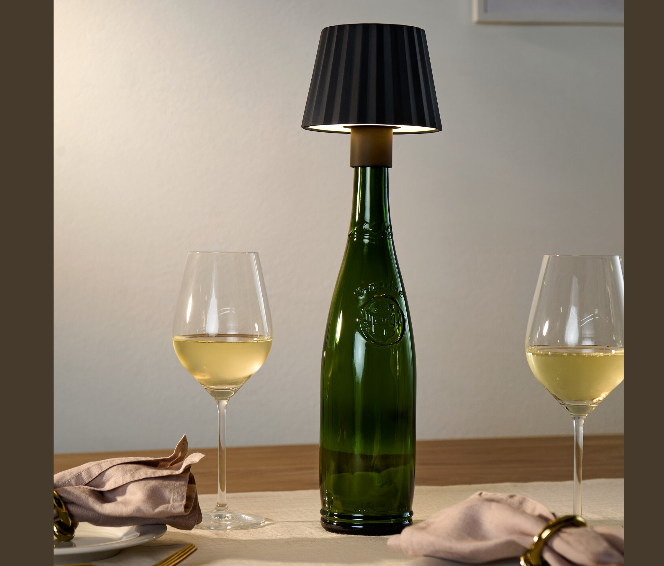 Egy asztalon egy zöld üveg fekete LED-es lámpa palackra termékkel, mellette két pohár fehérbor.