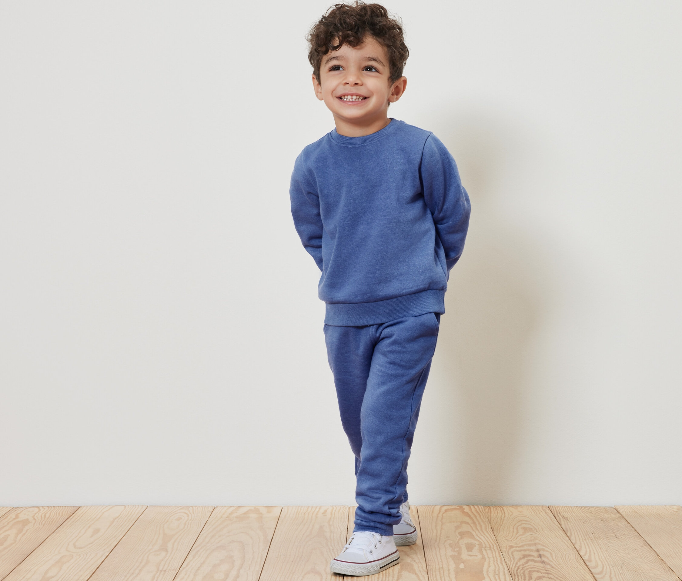 Egy mosolygó fiú világoskék tréningruhában és fehér tornacipőben pózol.