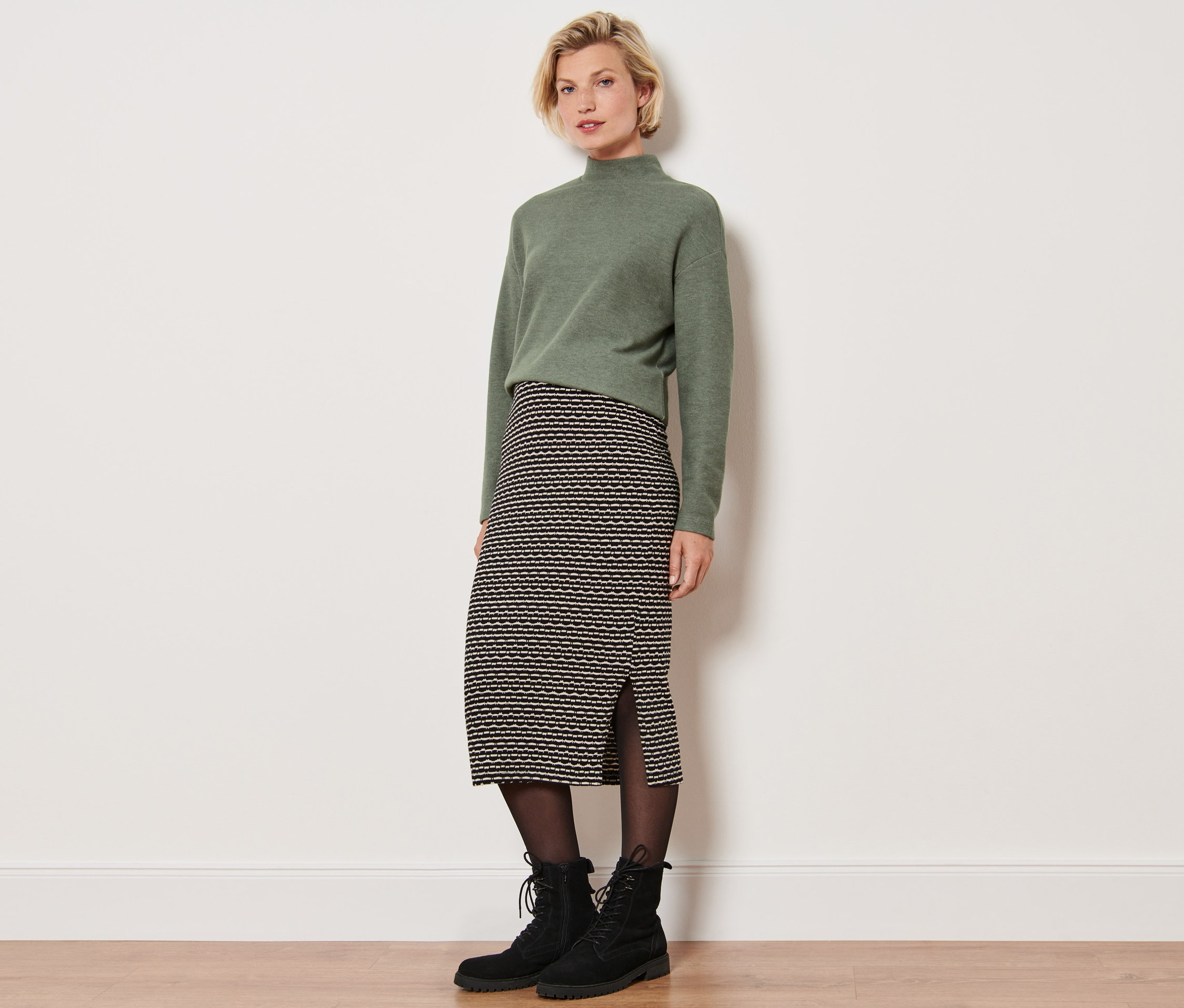 Egy nő áll a fal előtt, kötött szoknyát visel.