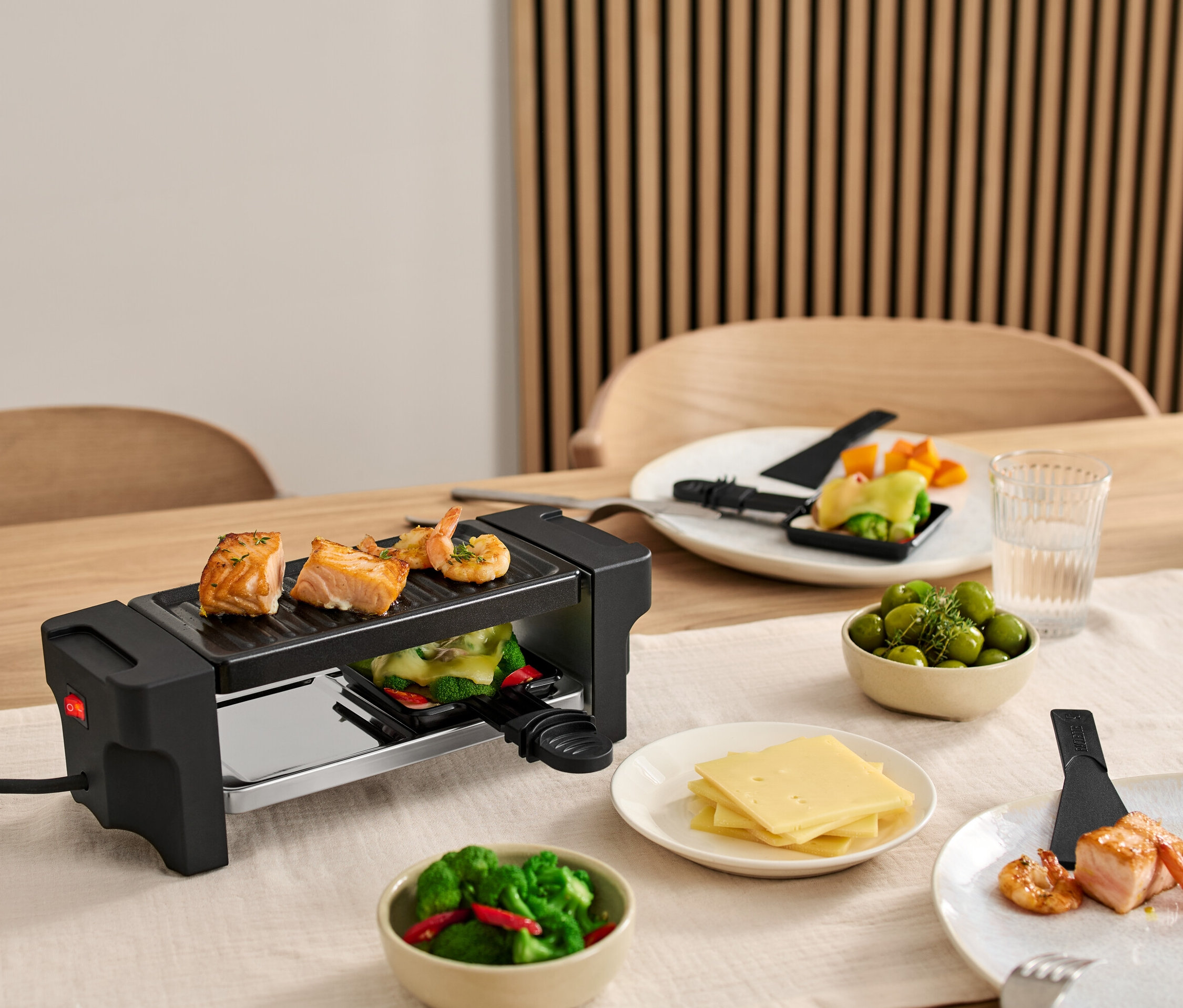 Egy kétszemélyes raclette grill van az asztalon, lazaccal és garnélával a grillen. Tányérok hozzávalókkal a grill körül.