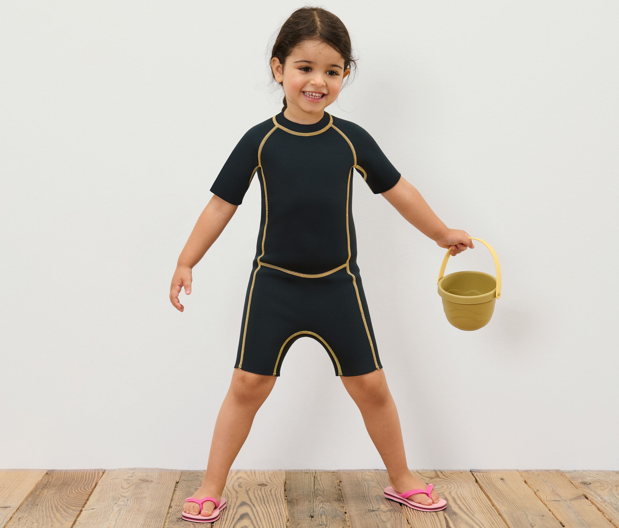 Kislány gyerek neoprénruhában sárga vödröt tart.