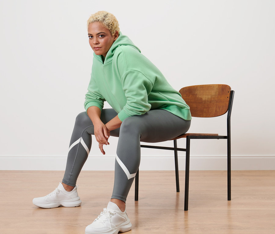 Egy nő ül egy széken, világoszöld kapucnis szabadidőfelsőt és szürke sportnadrágot visel.