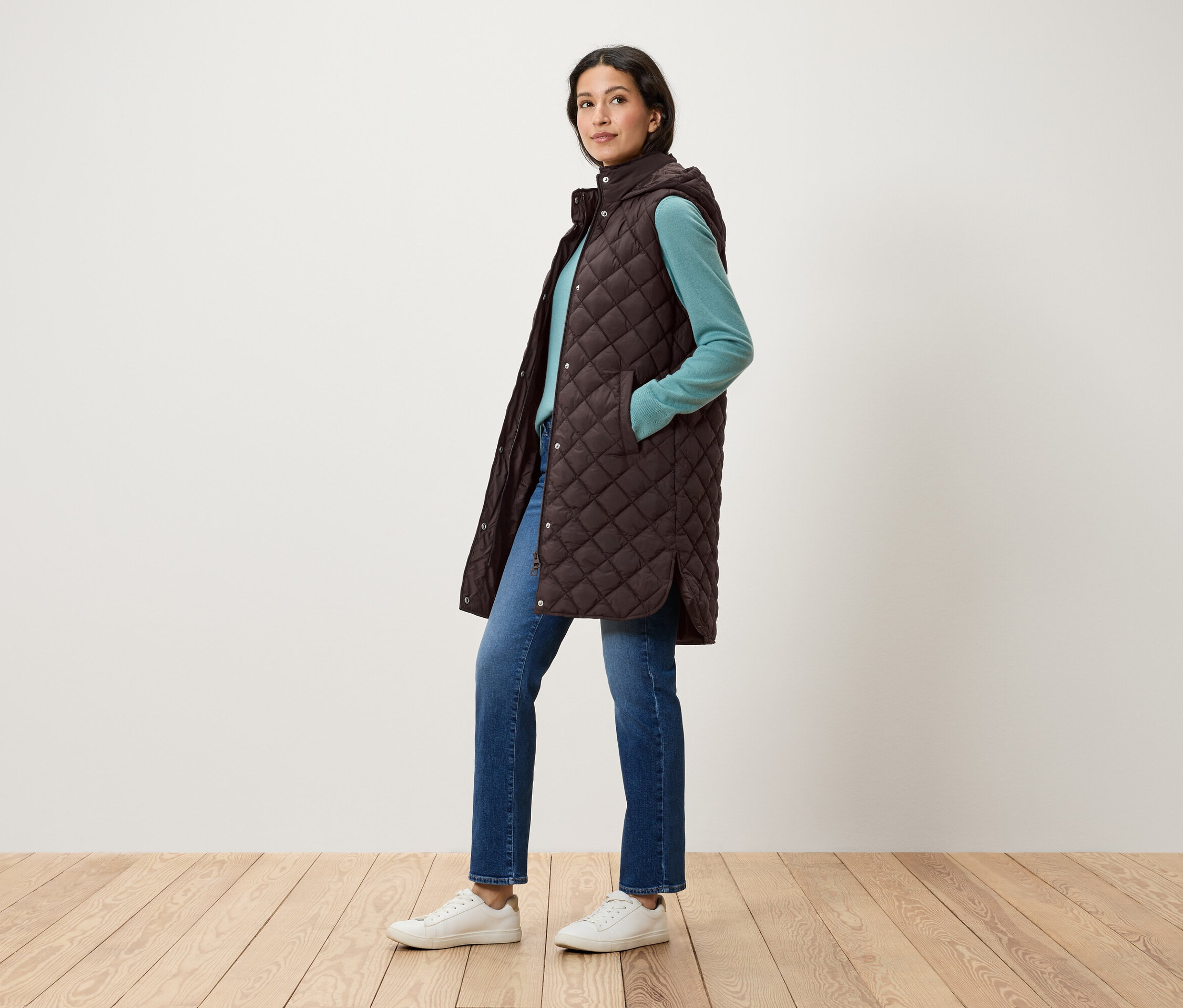 Nő pózol steppelt, barna, kapucnis mellényben, kék pulóverben, farmerban és fehér cipőben.