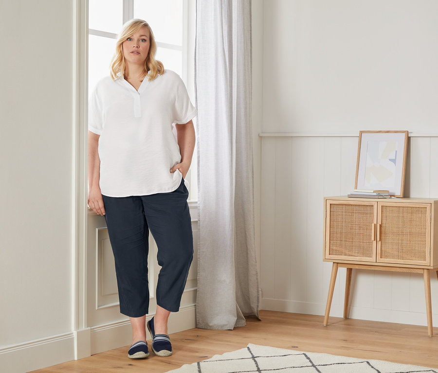 Egy nő egy ablak mellett pózol, oversize blúzban, lezser chino nadrágban és espadrillesben.