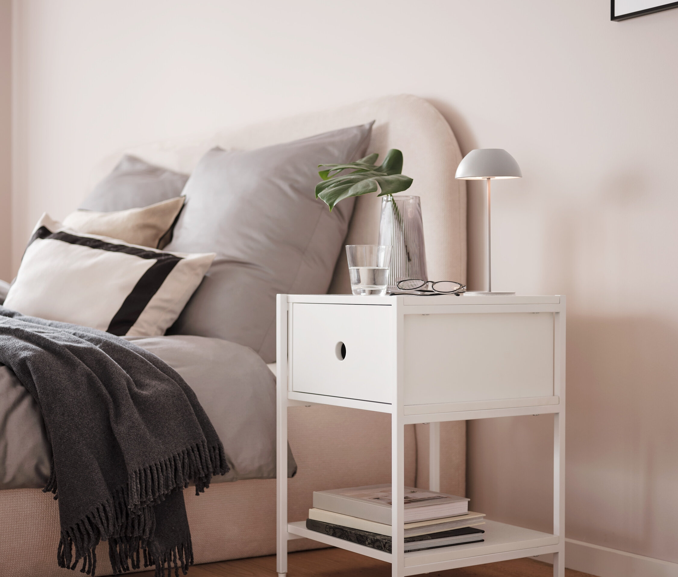 Szürke LED-es akkus asztali lámpa egy fehér éjjeliszekrényen, egy ágy mellett.