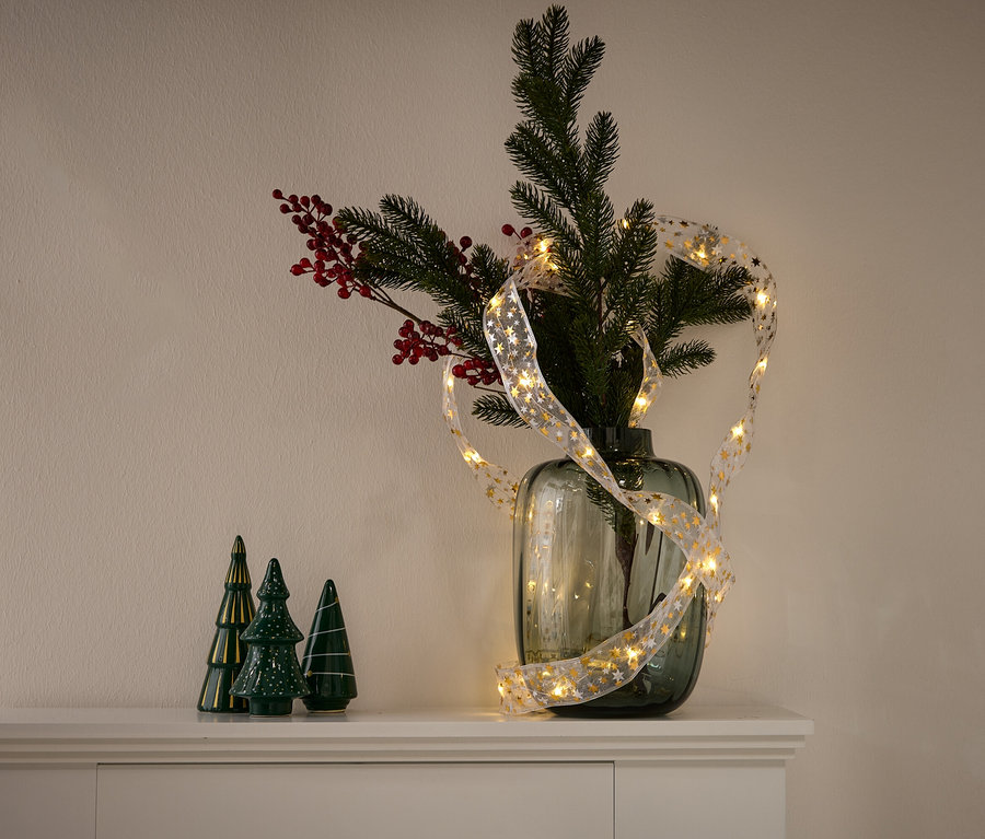 Egy váza fenyőágakkal, piros bogyókkal és egy aranyszínű csillagokkal díszített fényfüzérrel. Bal oldalon három zöld kerámia fenyőfa áll.