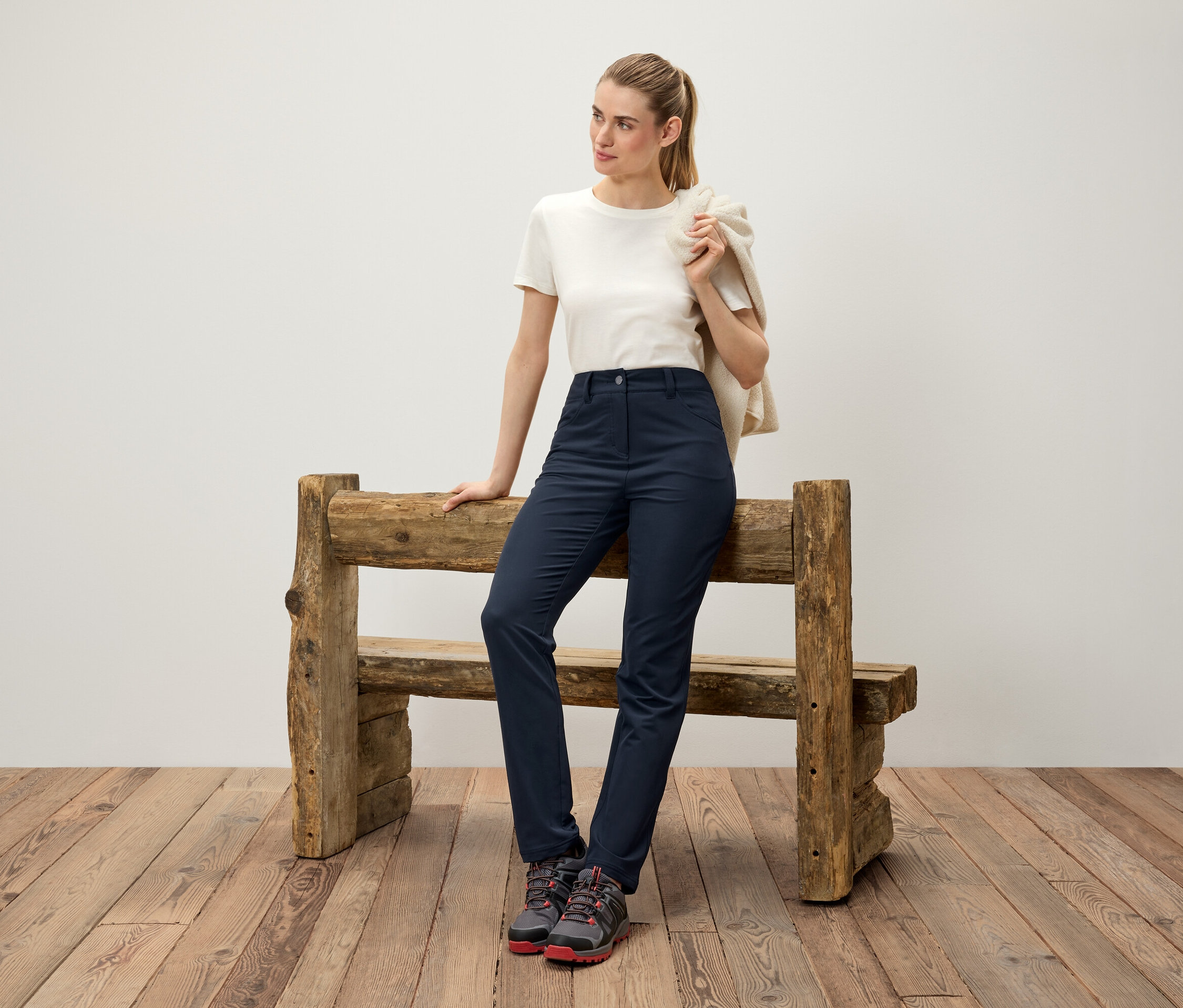 Egy nő egy fa padnak támaszkodik, krémszínű pulóvert visel a vállán, fehér pólót, sötétkék nadrágot és túrabakancsot.