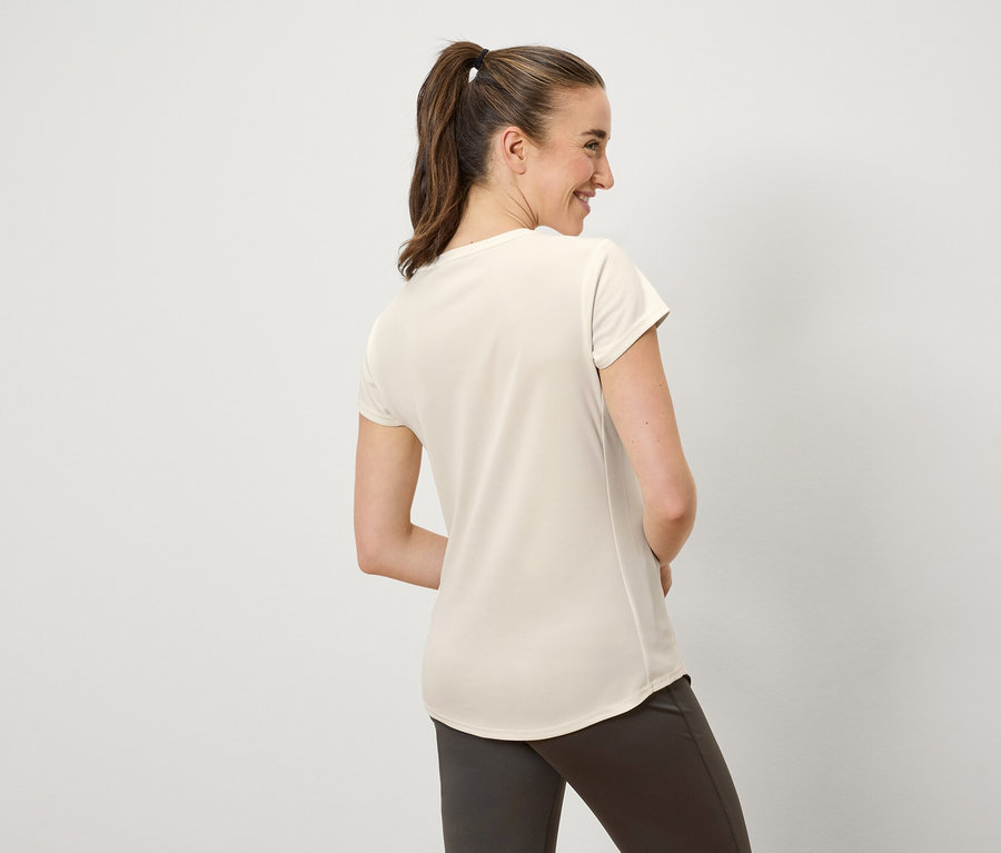 Egy barna hajú, lófarokba kötött nő mosolyog, és balra néz. Krémszínű pólót és barna nadrágot visel.