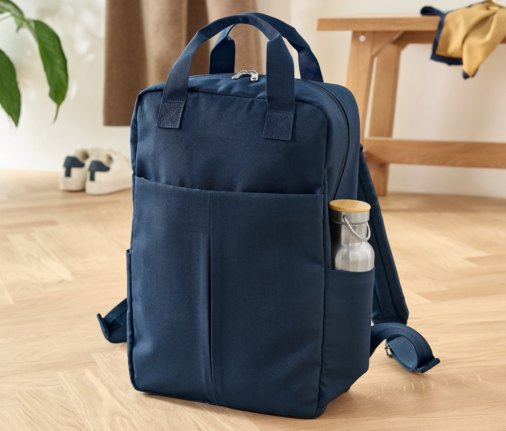 Egy sötétkék biztonsági hátizsák áll egy fa padlón. Az oldal zsebben egy vizes palack van.