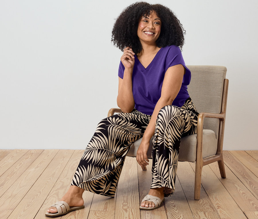 Nő ül a széken, lila, finom kötésű pulóvert és mintás palazzo nadrágot visel.