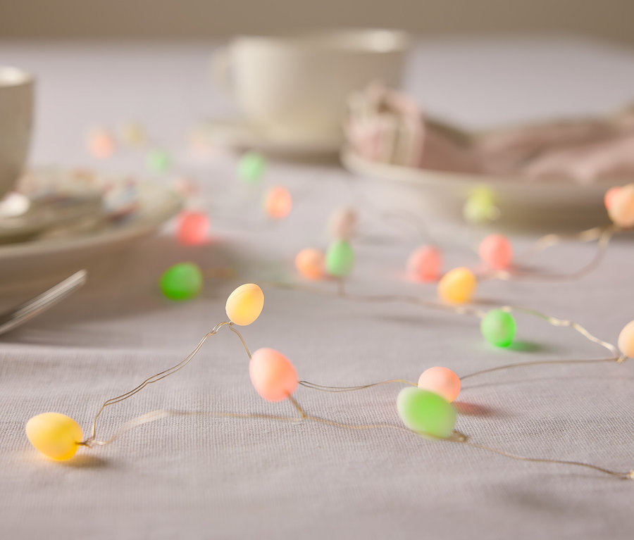 Közeli kép egy asztalról, amelyen edények és pasztell színű húsvéti tojás alakú fényfüzér található.