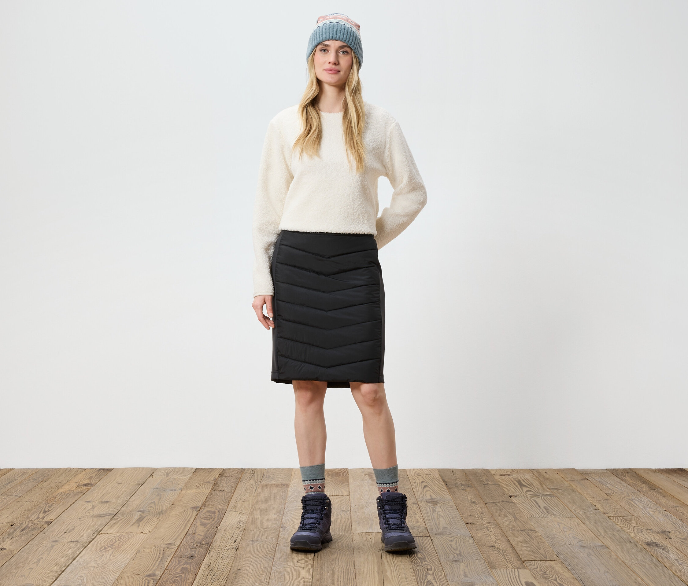 Nő áll a fapadlón, krémszínű pulóvert, fekete szoknyát, csizmát és kötött sapkát visel.