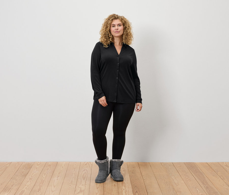 Egy szőke hajú nő áll fekete leggingsben, fekete hosszú ujjú ingben és szürke csizmában.