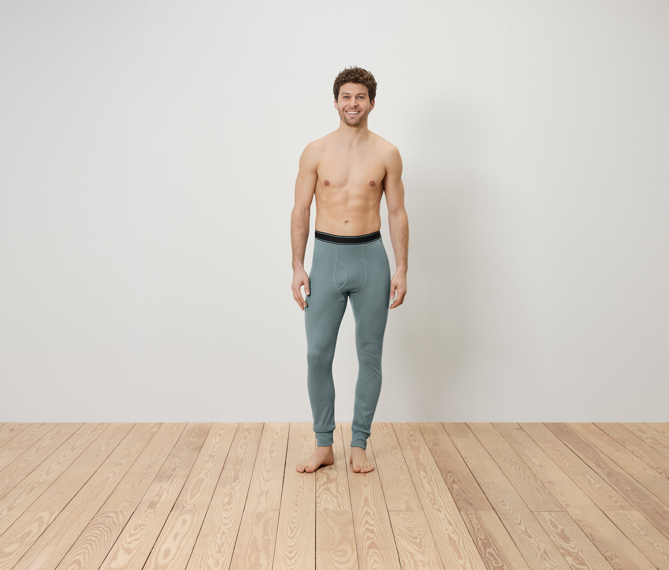 Egy felsőtest nélküli férfi áll egy fapadlón, szürke leggingset visel.