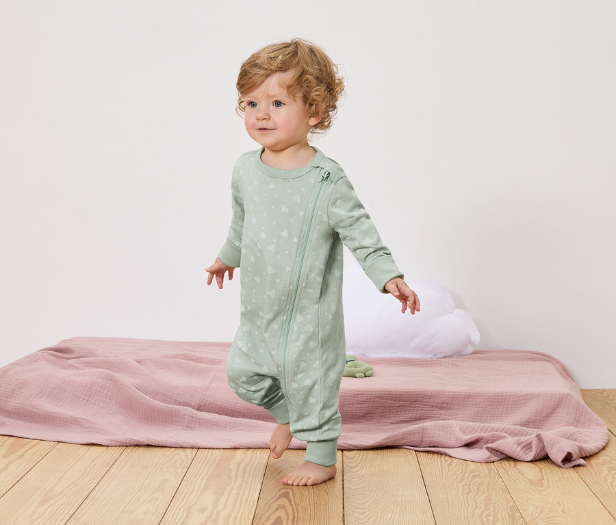 Egy göndör szőke hajú kisgyermek mezítláb sétál egy fapadlón. Világoszöld, cipzáras, szívecskés mintás pizsamát visel.