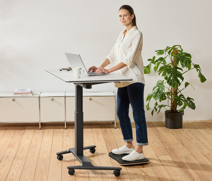 Egy nő áll egy állítható magasságú asztalnál és laptopot használ, mellette egy bögre áll.