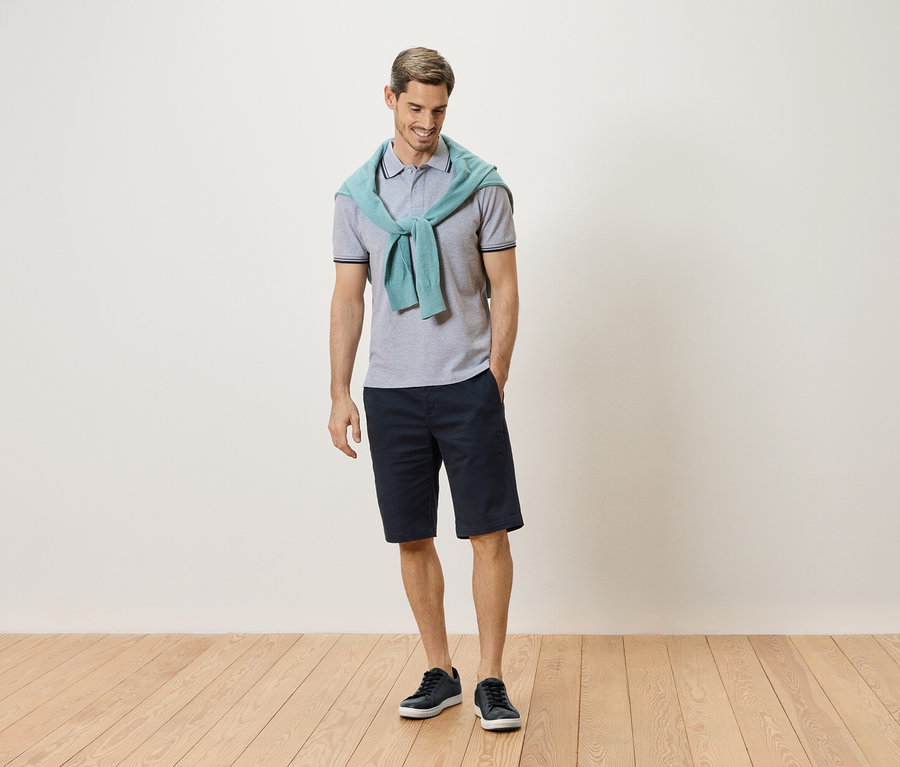 Férfi áll világoskék pulóverrel a nyakában, szürke pólóinggel, sötétkék rövidnadrággal és kék tornacipővel.