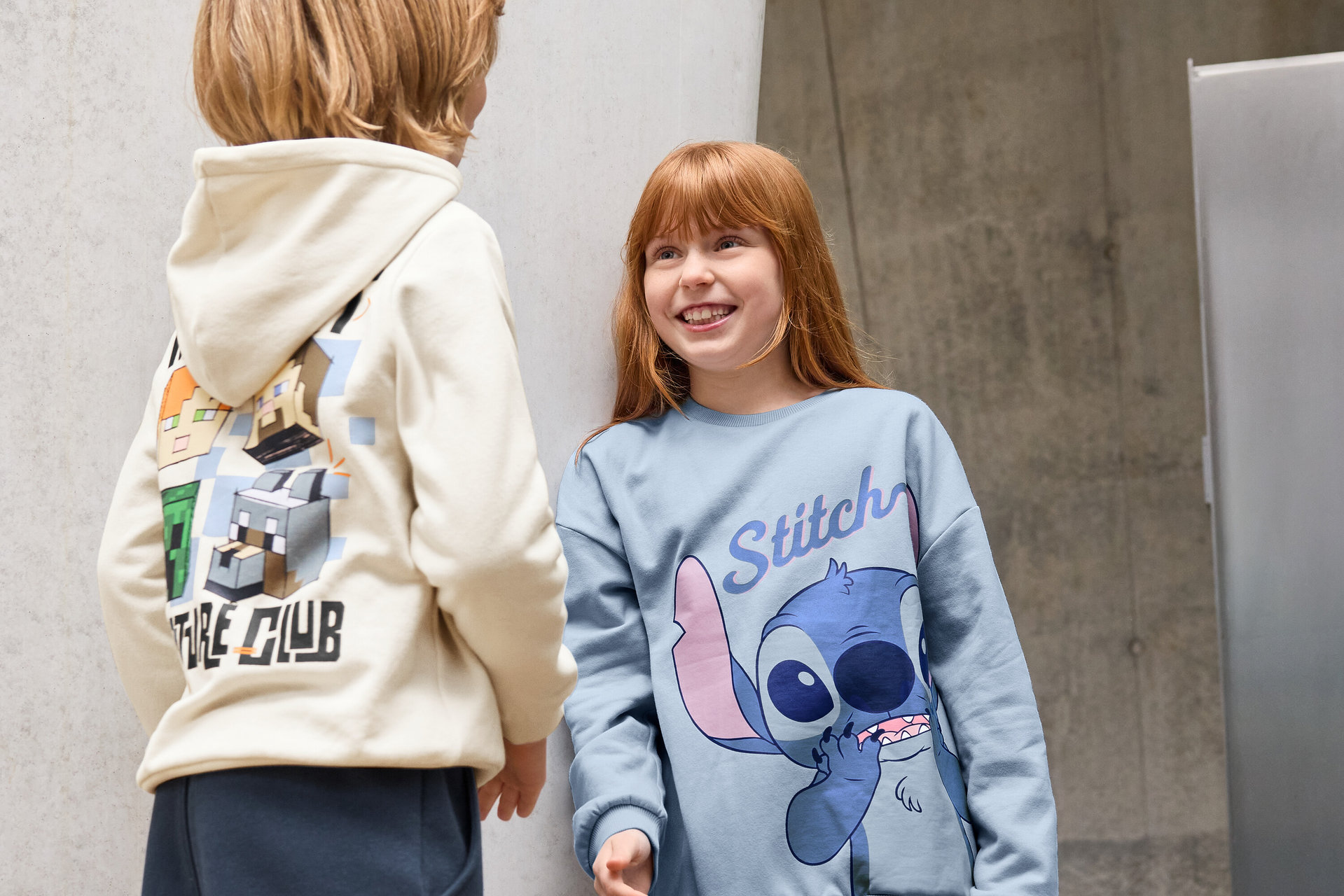 Két gyerek áll egy fal előtt. Az egyik gyerek Kinder-Jogginghose-t és egy Minecraft figurákkal ellátott kapucnis pulóvert visel. A másik gyerek 3 Kinder-Leggings-et, schwarz-ot és egy Stitch pólót visel.