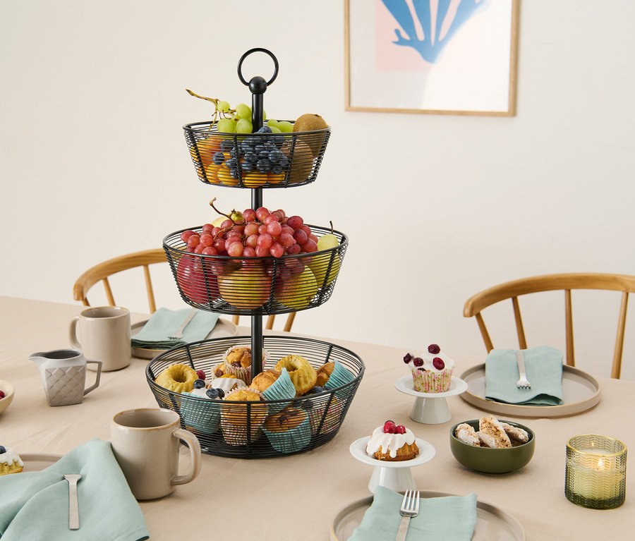 Egy háromszintes, fémhuzalból készült gyümölcskosár áll az asztalon süteményekkel, gyümölcsökkel és edényekkel.