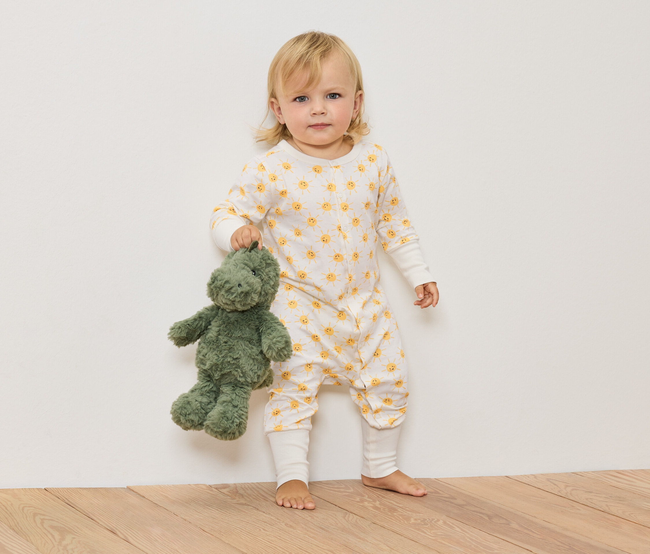 Egy szőke gyermek napocska mintás overallban zöld plüss dinoszauruszt tart.