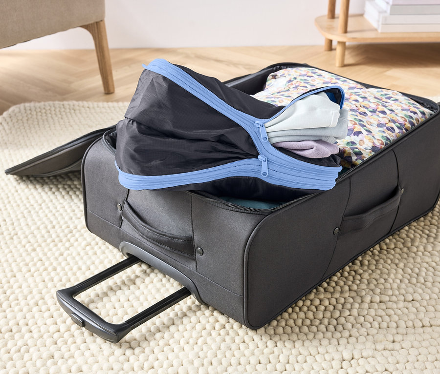 Egy nyitott, szürke bőrönd egy szőnyegen, ruhákkal és egy kék cipzáras fekete csomagoló kockával megtöltve.