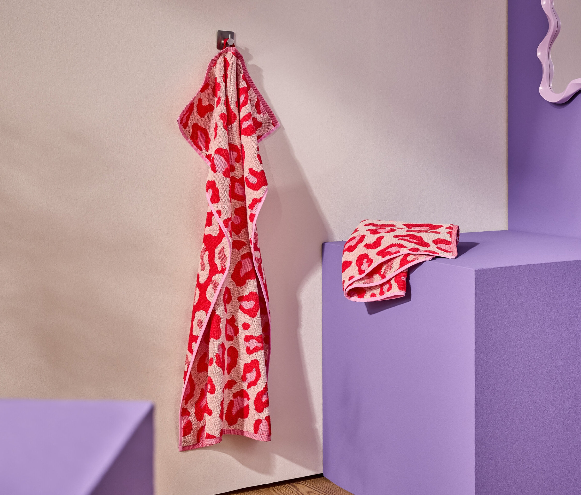 Egy vörös-rózsaszín, leopárdmintás törölköző lóg egy kampón. Egy összehajtogatott törölköző egy lila talapzaton fekszik.