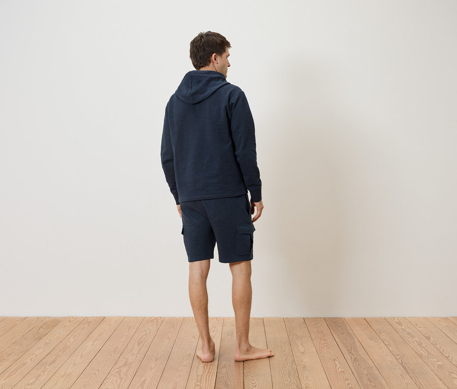 Hátulról látható férfi sötétkék kapucnis pulóverben és rövidnadrágban, fa padlón.