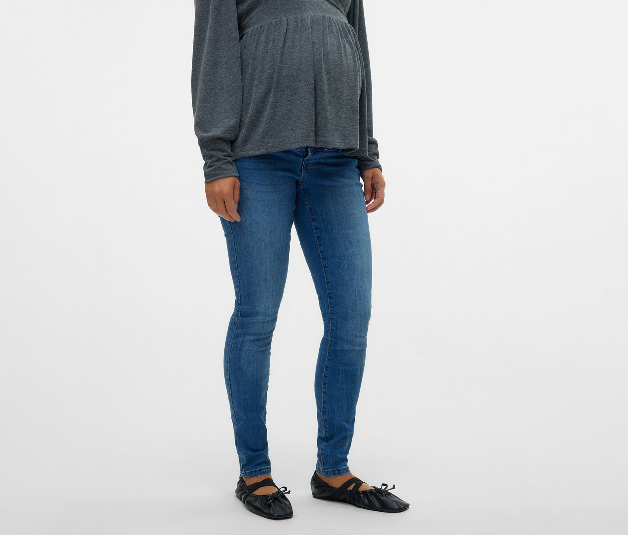 Egy várandós nő szürke felsőt és MAMALICIOUS kismama farmernadrágot visel fekete balerinacipővel.