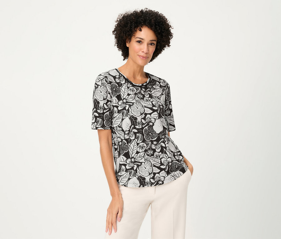 A göndör hajú nő virágmintás Olsen pólót és világos nadrágot visel.