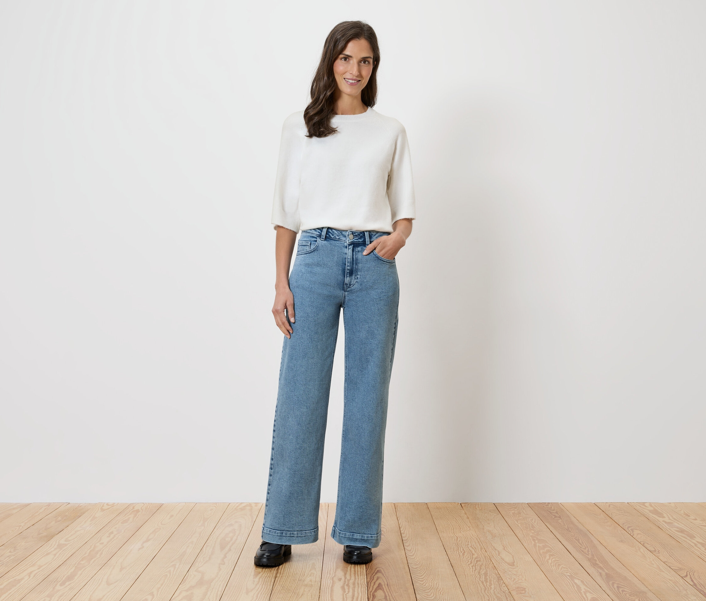 Egy nő áll egy fehér fal előtt, fehér blúzt és bő farmert visel.