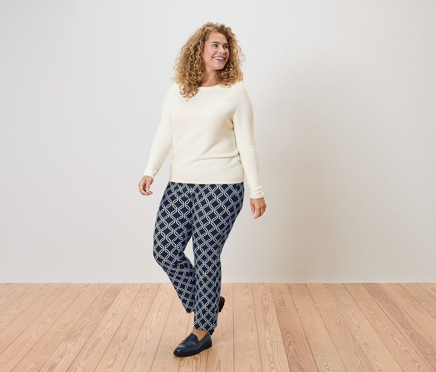 Göndör hajú nő, krémszínű pulóverben és geometrikus mintás sötétkék nadrágban.