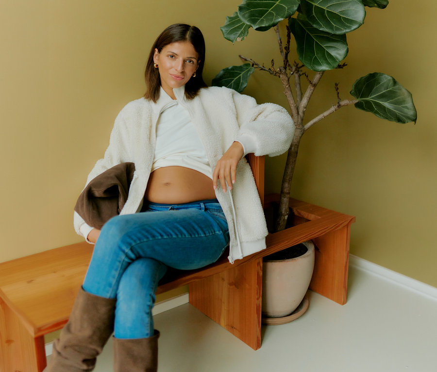 Egy terhes nő ül egy padon egy növény mellett, és egy fehér póló és farmer felett egy nyitott MAMALICIOUS 3in1 kismama plüssdzsekit visel.